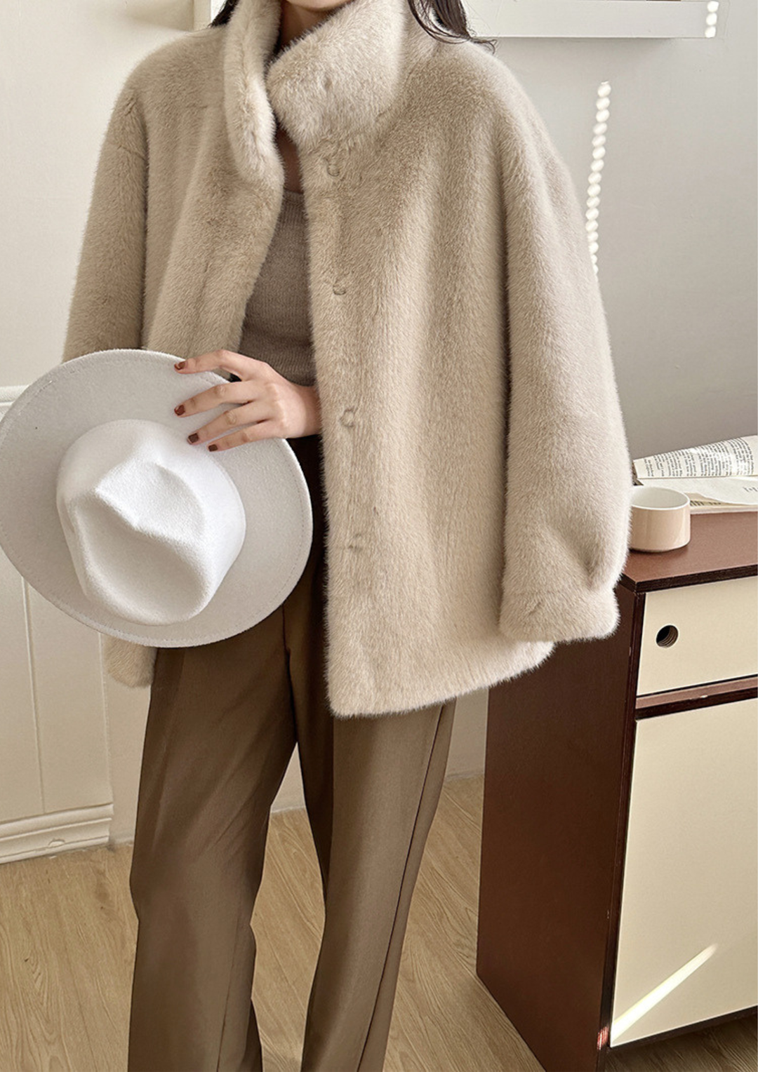 lily Mila plush mink fur coat in beige with a stand collar, worn with brown trousers and holding a white hat.